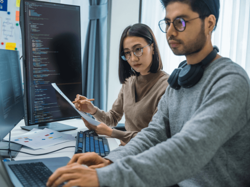 Two people working at a desk with computer monitors displaying code.