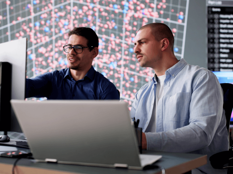 Two men working at a desk with computer monitors, with a data visualization displayed in the background.