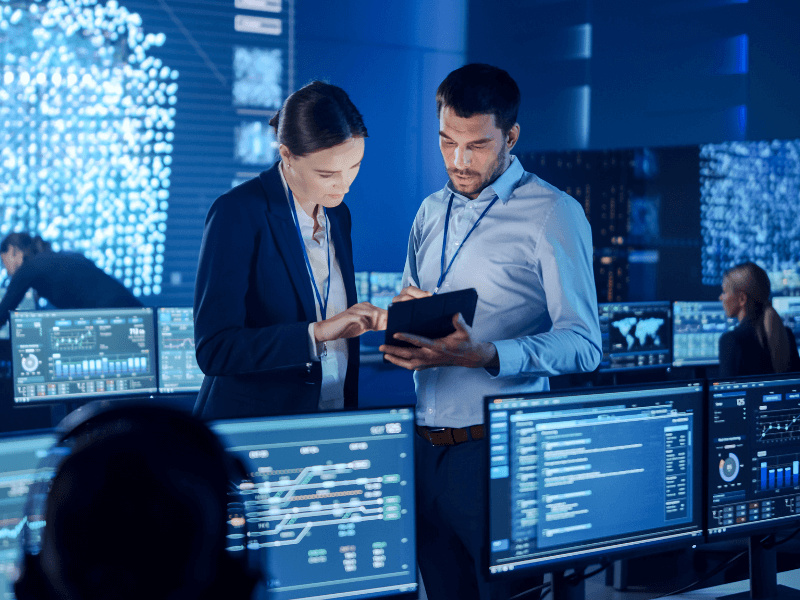 Two professionals examining a tablet in a high-tech control room filled with screens displaying data.