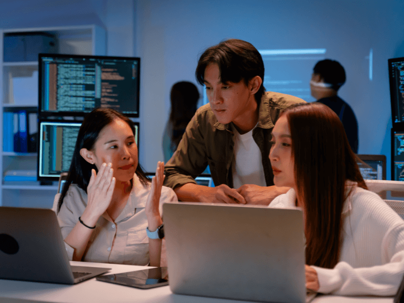 Three people collaborating in a tech environment with laptops and computer screens displaying code.