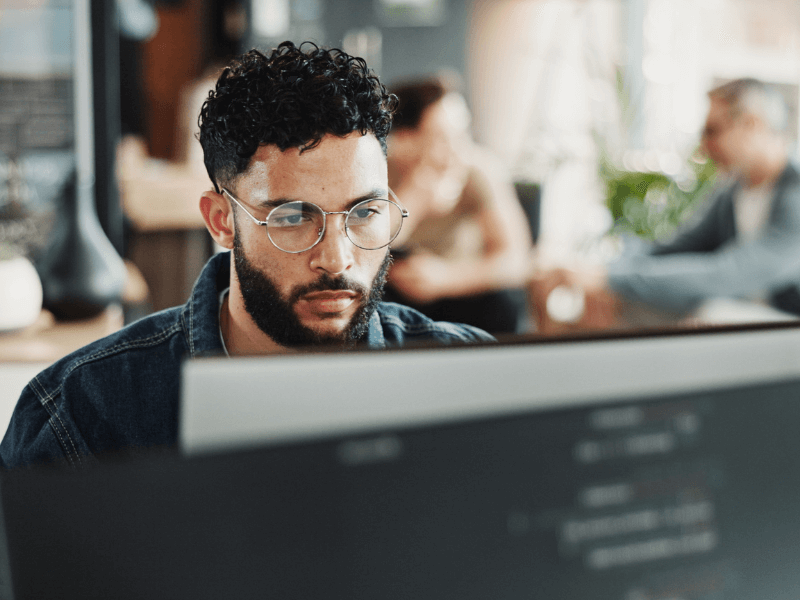 A person with curly hair and a beard focuses on a computer screen in an office setting.