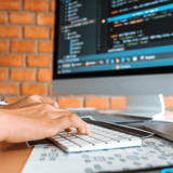 Hands typing on a keyboard in front of a monitor displaying code, with a brick wall in the background.