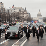 A motorcade in a city with a prominent building, likely the US Capitol, in the background.