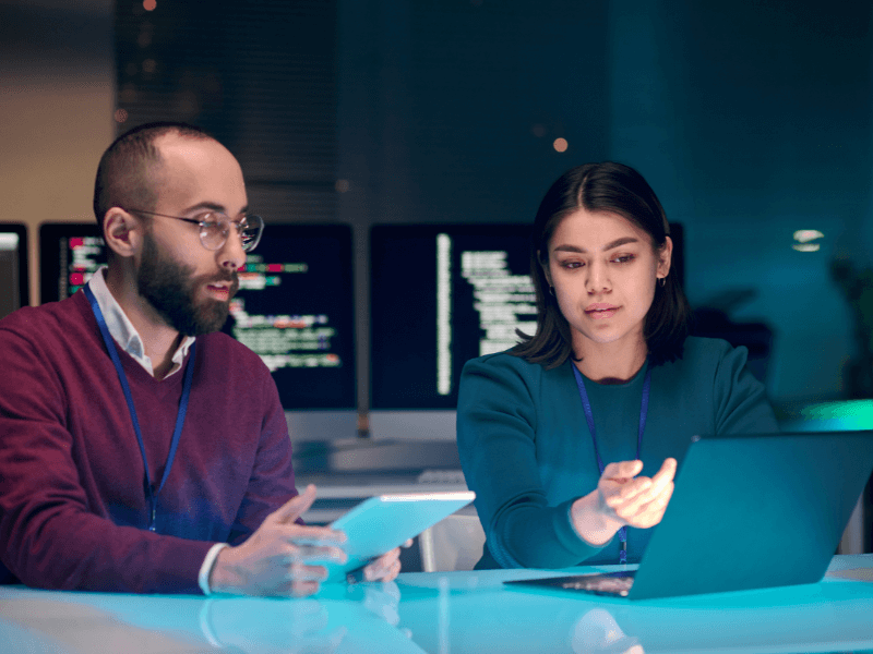 Two people working on a tablet and laptop in a dimly lit office with code on monitors in the background.