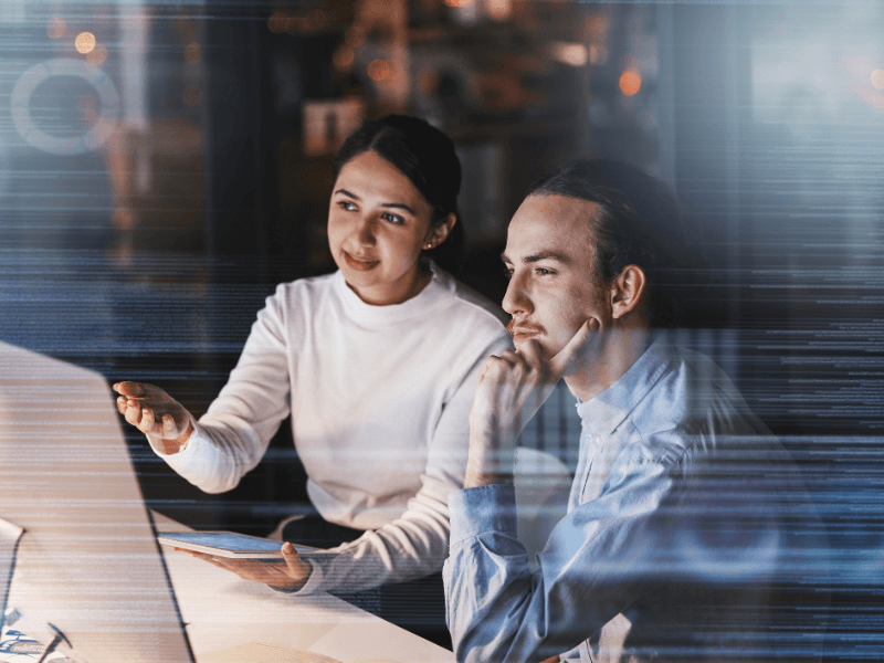 Two people at a computer in an office setting, engaged in discussion.