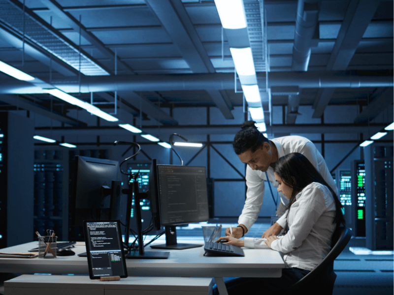 Two people collaborating at a desk in a tech environment, surrounded by monitors and servers.