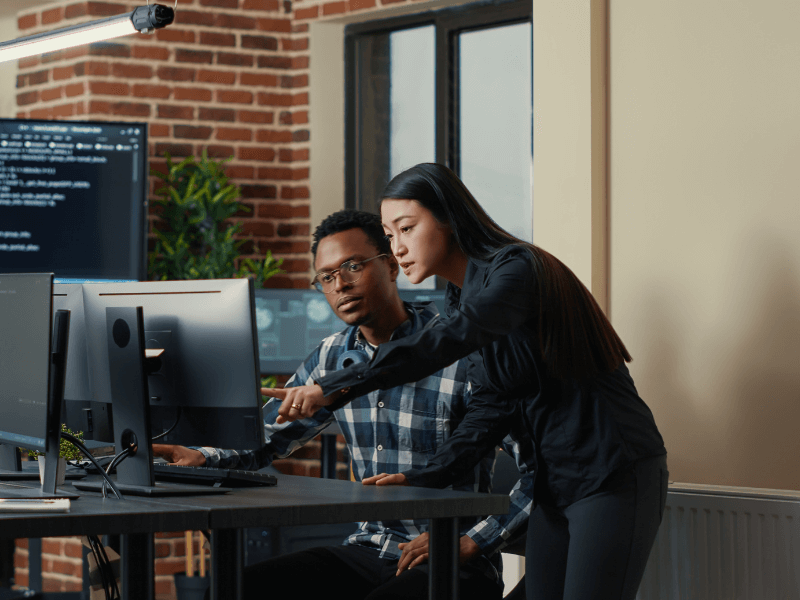 Two people collaborating at a desk with computers in an office