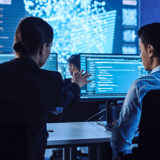 Two people in a tech office analyzing data on computer screens.