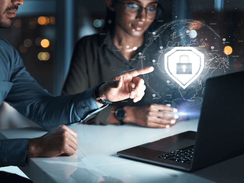 Two people in an office observe a holographic padlock and shield symbol, representing cybersecurity