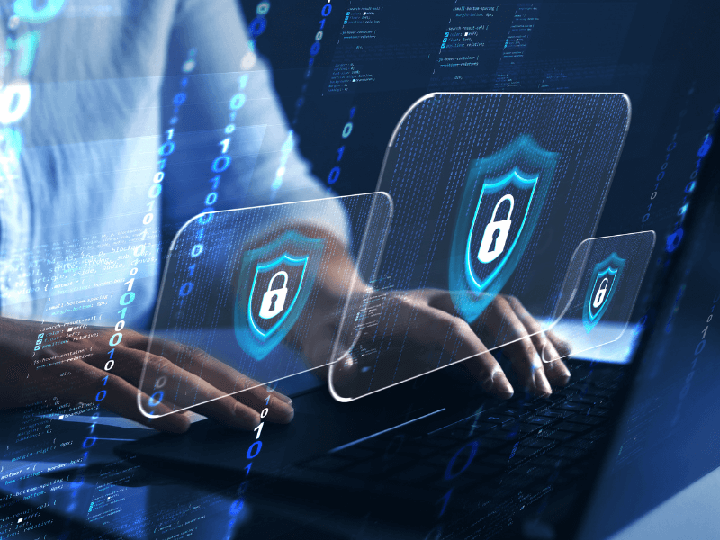 Person typing on a laptop with holographic security shields and digital code overlay.