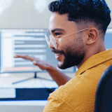 A person in a mustard-yellow shirt gesturing in front of a computer monitor displaying blurred code.