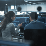 Two people discussing code at a computer workstation in an office setting.