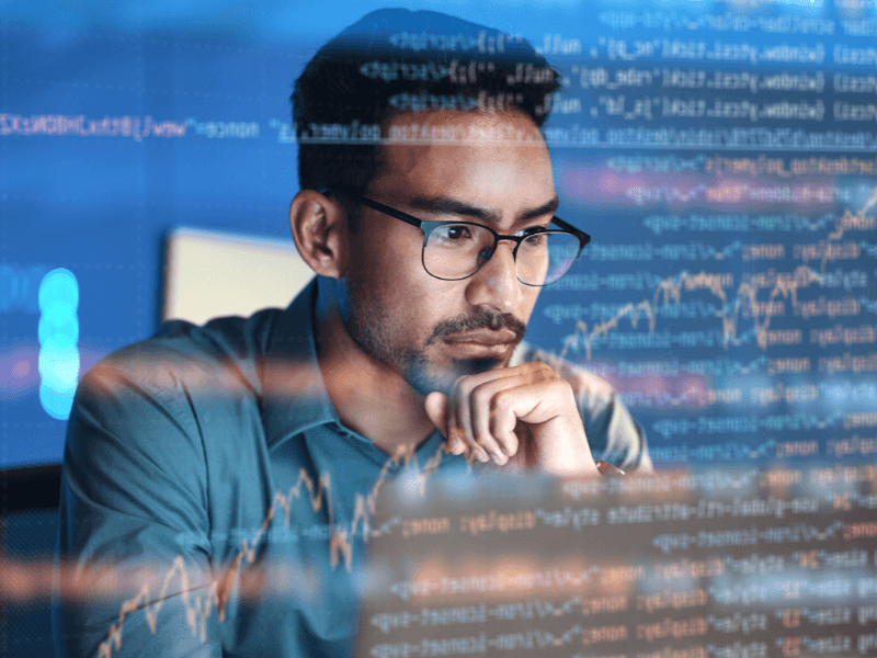 A man with glasses focuses on a computer screen, with an overlay of code and stock market graphs